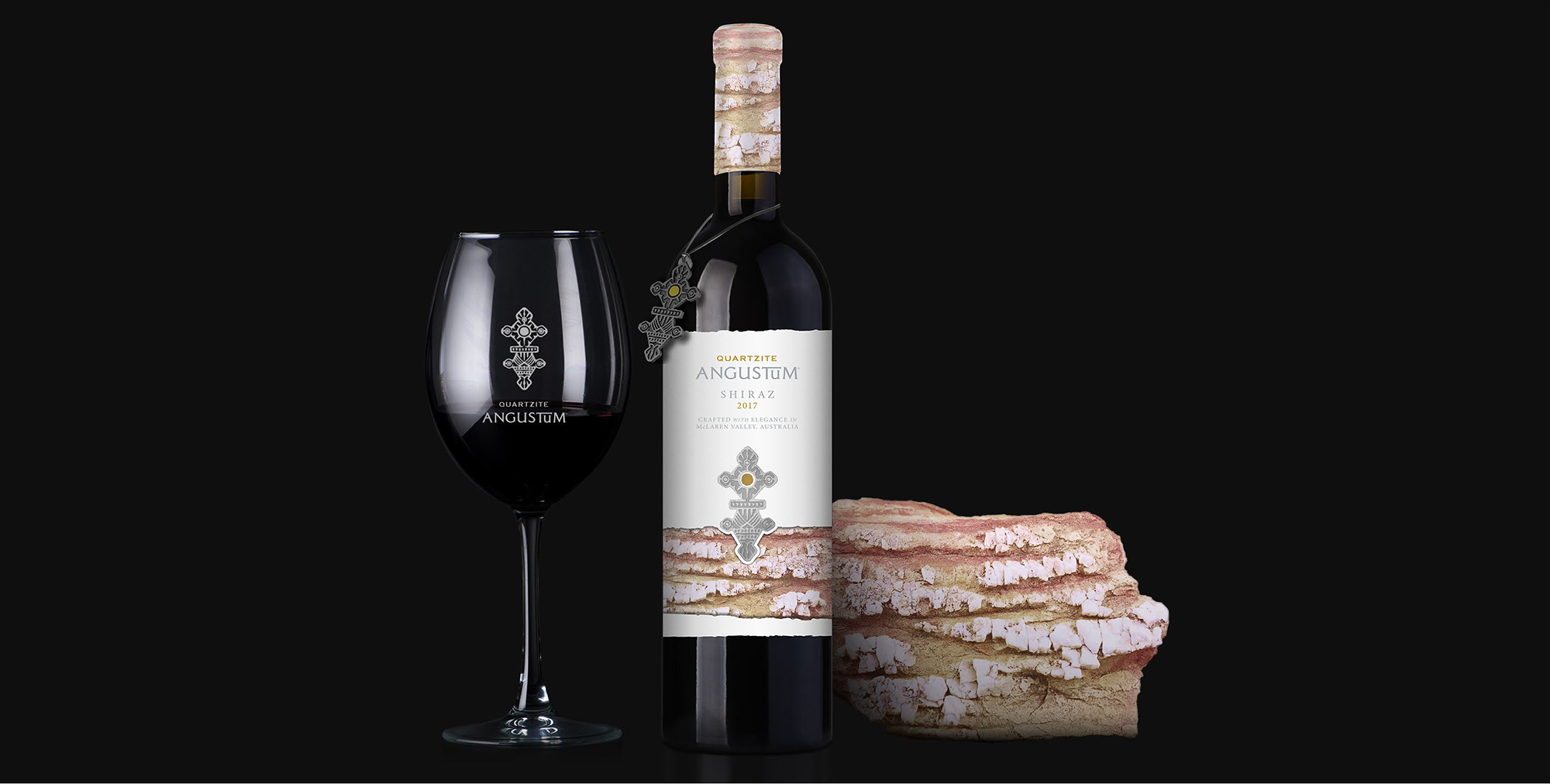 A bottle of red wine with a cork and a wine glass, alongside a stack of cheese wedges, all set against a solid black background. The bottle label and the glass are printed with a silver cross design.